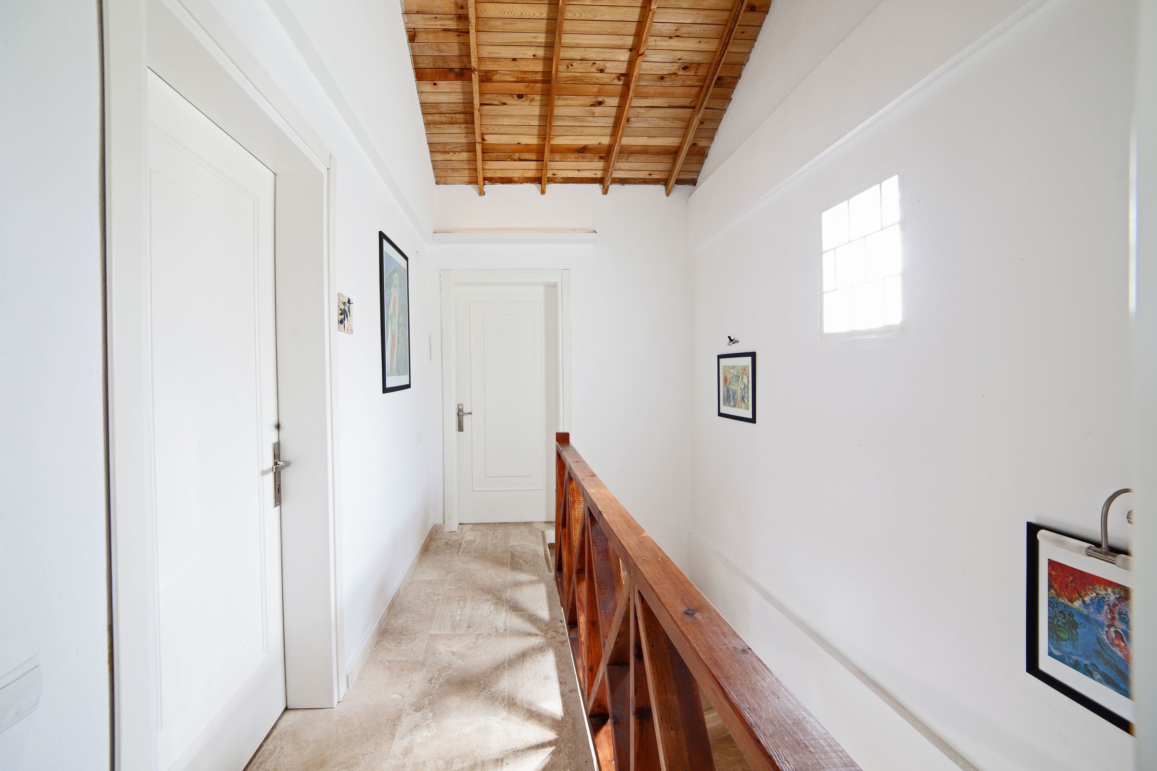 A hallway with white walls, a wooden ceiling, and a wooden bench. There are doors and framed pictures on the walls.