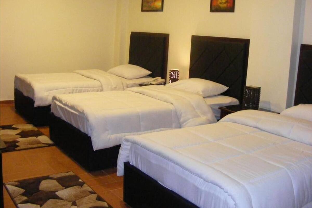 Ein Hotelzimmer mit vier Einzelbetten, jeweils mit dunklem Kopfteil und weißer Bettwäsche. Darüber hängen zwei an der Wand befestigte Bilder.