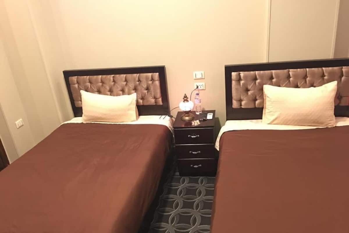 Ein Hotelzimmer mit zwei Betten, jeweils mit Kopfstütze, einem Nachttisch mit Lampe und einer kleinen Kommode.