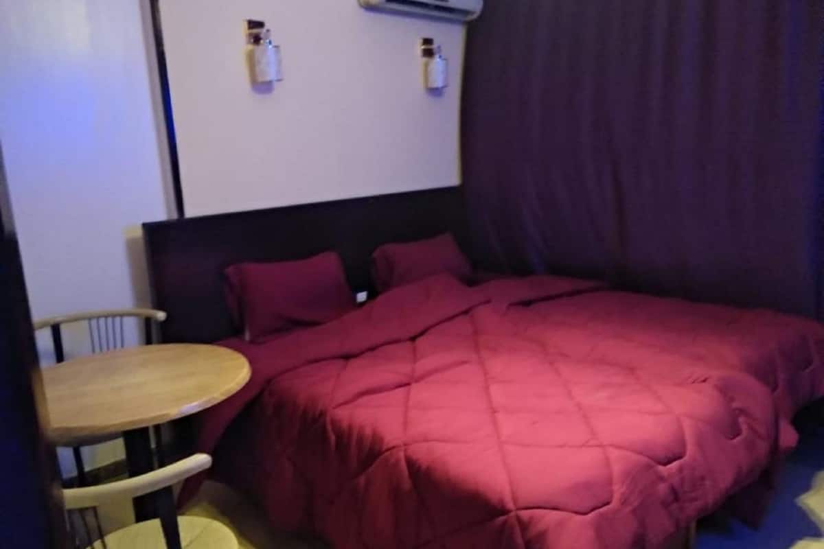 Ein kleiner, kompakter Raum mit einem Bett, einem runden Tisch und zwei Stühlen. Über dem Bett befindet sich eine wandmontierte Klimaanlage.