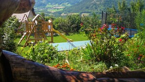 A playground with a slide and climbing structure, surrounded by lush greenery and a scenic mountain view.