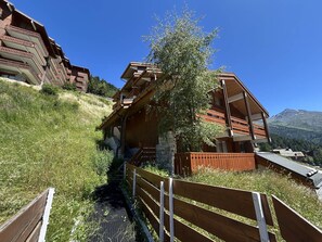 Una cabaña de madera con escaleras de piedra, rodeada de vegetación exuberante y un paisaje montañoso.