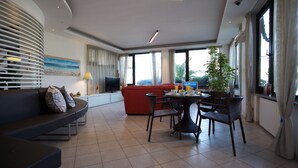 A modern living room with a curved sofa, a dining area with a round table and chairs, and large windows offering a view of the outdoors.