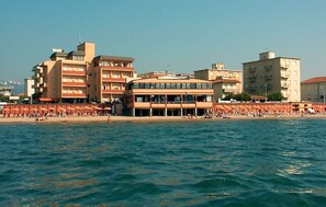 Ein Hotel an der Küste mit mehreren Etagen, ein Strand im Vordergrund und einige Gebäude im Hintergrund.
