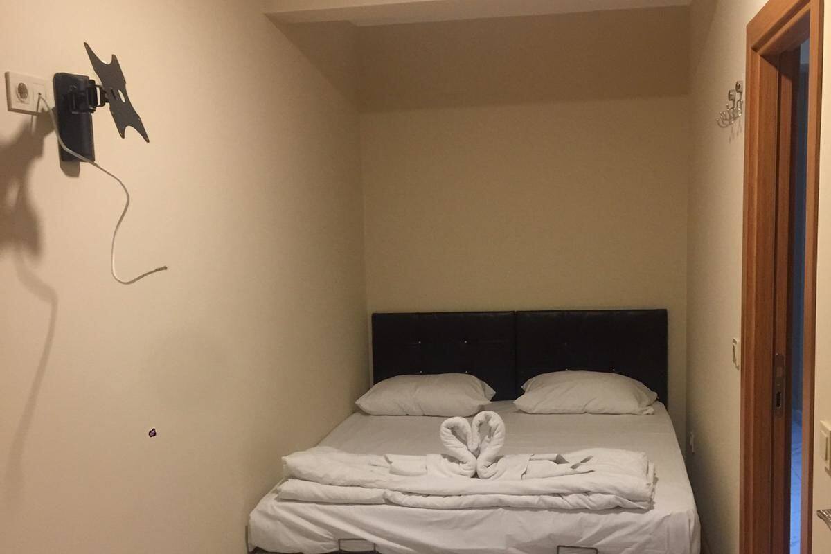 Uma cama pequena e bem arrumada, com cabeceira, luminária fixada na parede e uma porta à direita.
