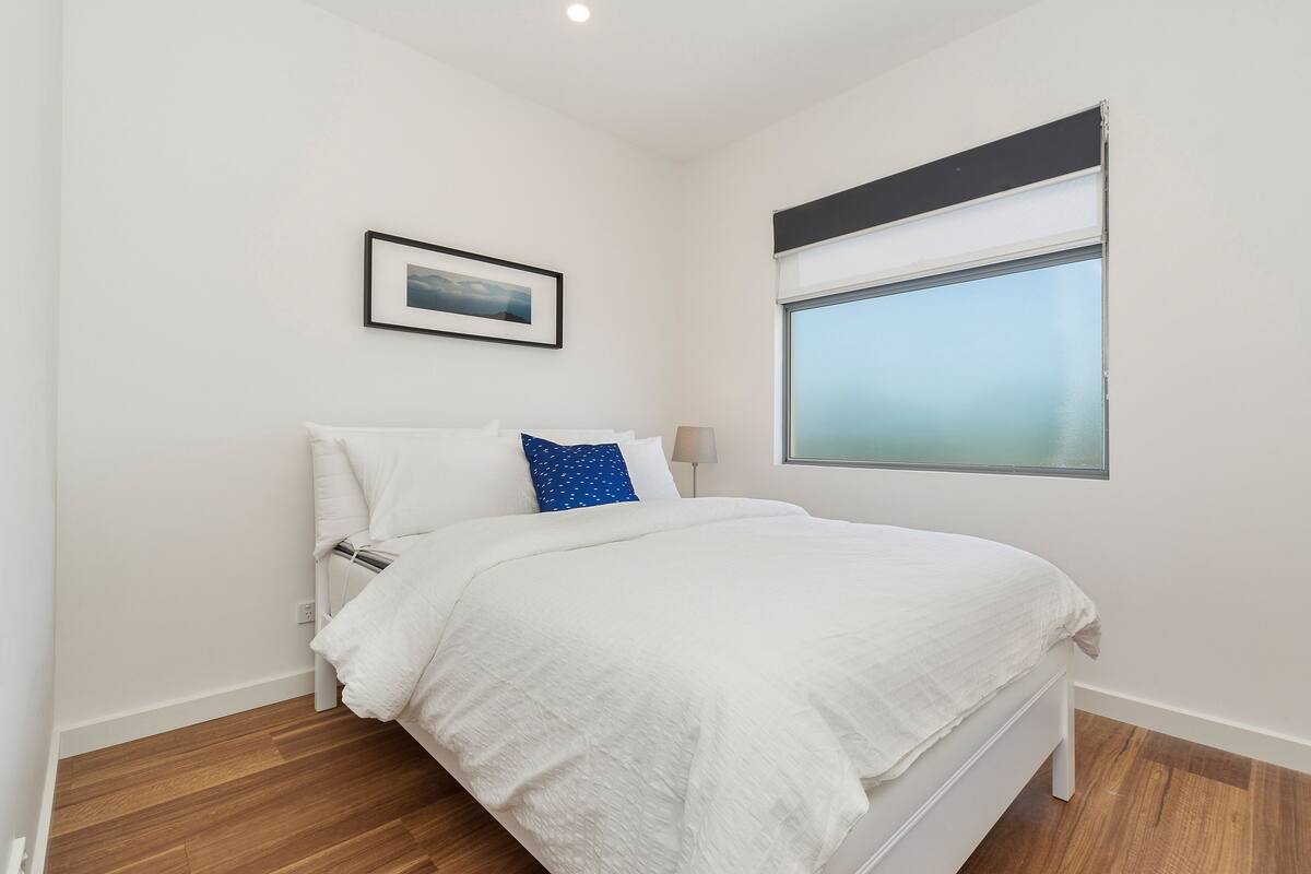 A bedroom with a bed, white walls, a framed picture, a window, and a wooden floor.