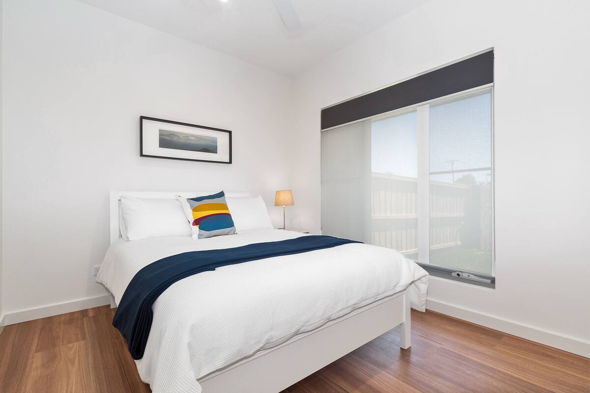 A bedroom with a bed, a framed picture on the wall, a window, and a nightstand with a lamp.