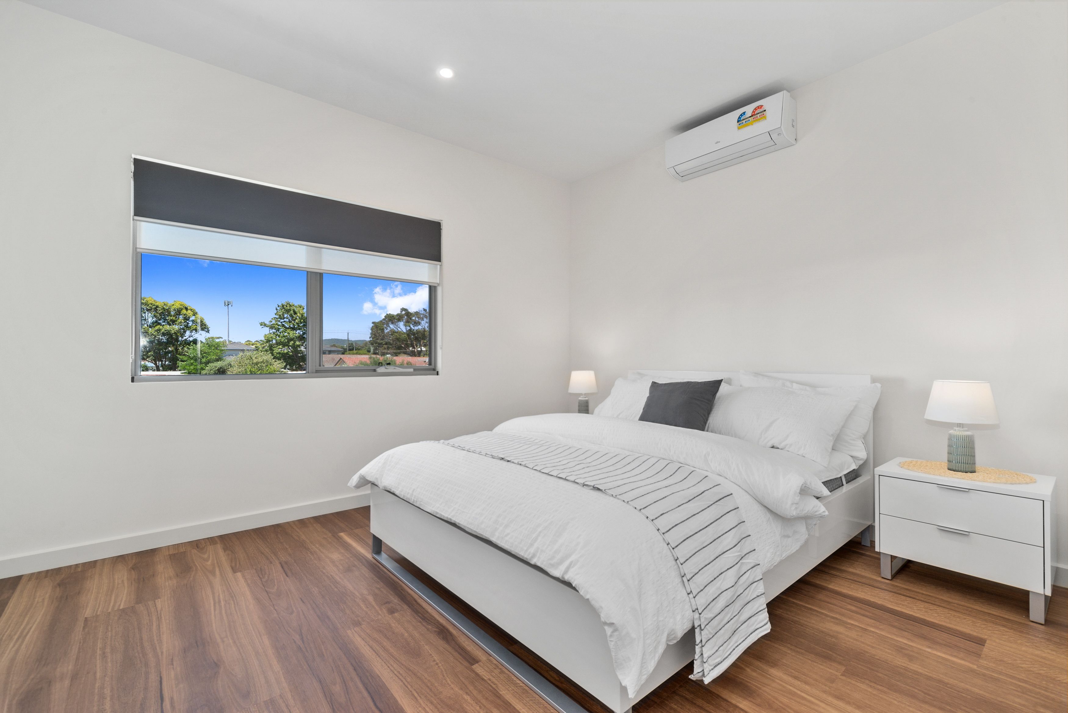 A bedroom with a large window, a bed, a bedside table, and an air conditioning unit.