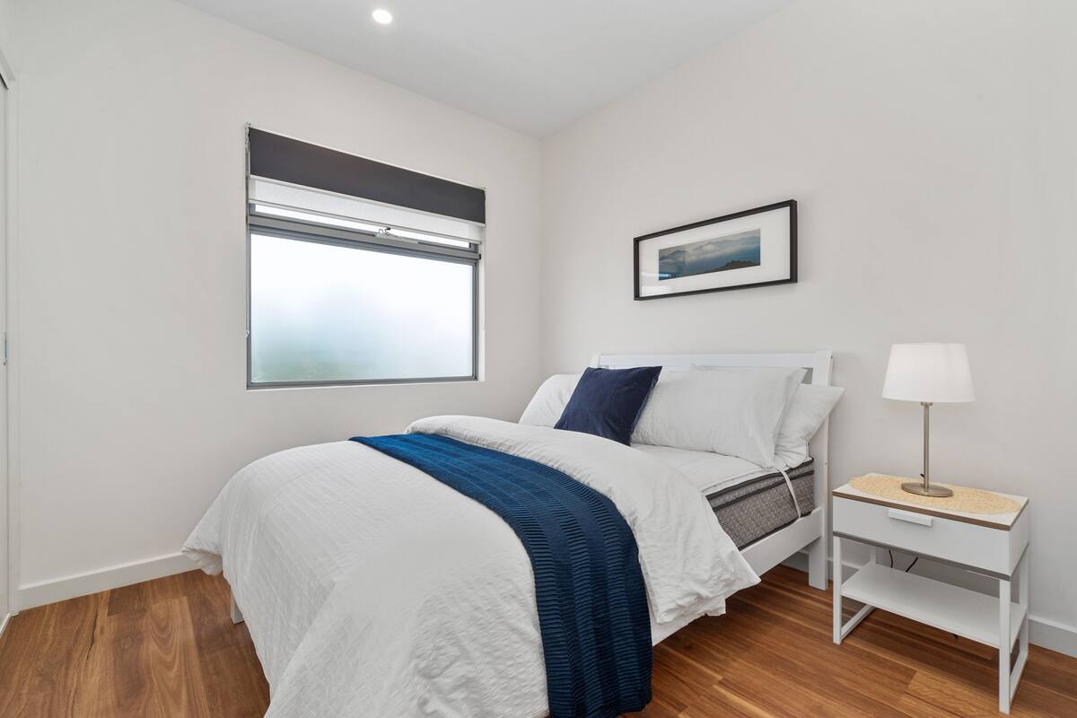 A modern bedroom with a bed, a bedside table with a lamp, a window with blinds, and a framed picture on the wall.