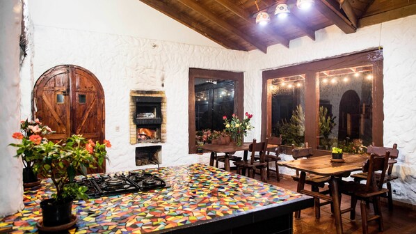 Un coin repas rustique avec une table recouverte de mosaïques, des chaises en bois, une cheminée et une grande porte en bois.