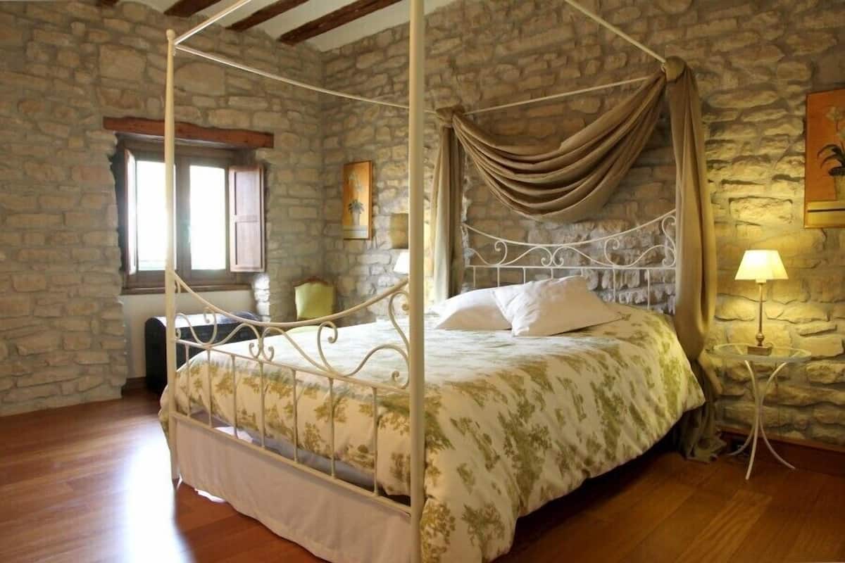 Ein Bett mit Baldachin, eine Steinwand, ein Fenster mit Holzläden und ein kleiner Tisch mit Lampe.