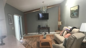 A living room with a wooden floor, a beige sofa, a coffee table, a TV mounted on the wall, and a painting on the wall. - 3 bedroom Pool home. Quiet Neighbourhood (Spring hill)