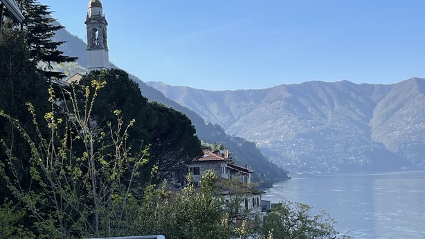 A lakeside view with a bell tower, a stone building, and a metal railing. - Lake Front with Large Balcony, Lake Access & Boat Dock (NESSO)