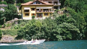 A yellow house with a balcony, surrounded by greenery and a body of water. - Lake Front with Large Balcony, Lake Access & Boat Dock (NESSO)