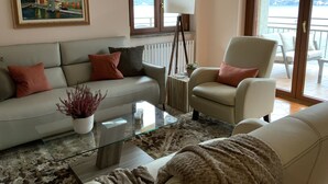 A living room with a sofa, armchair, and a glass coffee table. There is a painting on the wall and a view of the outdoors through a window. - Lake Front with Large Balcony, Lake Access & Boat Dock (NESSO)