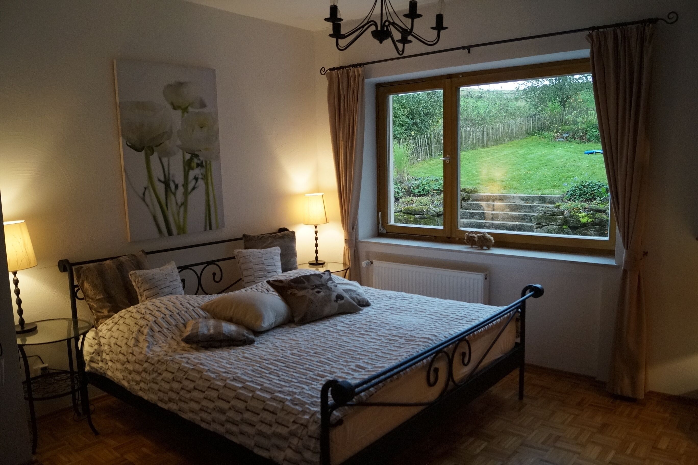 Ein Schlafzimmer mit einem Bett, Nachttischlampen, einem Fenster mit Vorhängen, einer Heizung und einem Bild an der Wand.