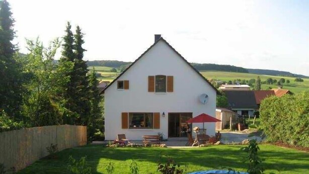 Une maison à deux étages, avec une façade blanche, un parasol rouge et un trampoline dans le jardin.