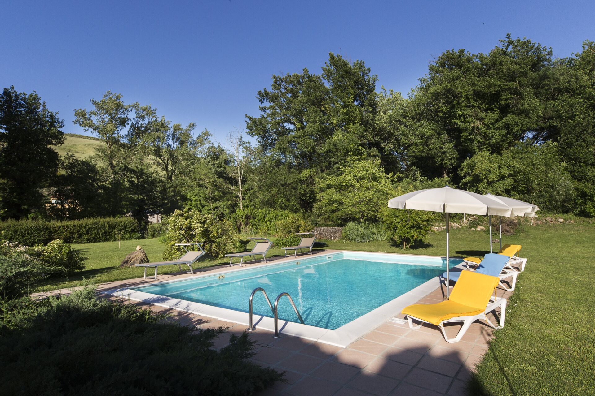 A rectangular swimming pool with a clear blue water, surrounded by a tiled deck. There are lounge chairs with yellow and white cushions, and a white umbrella providing shade.