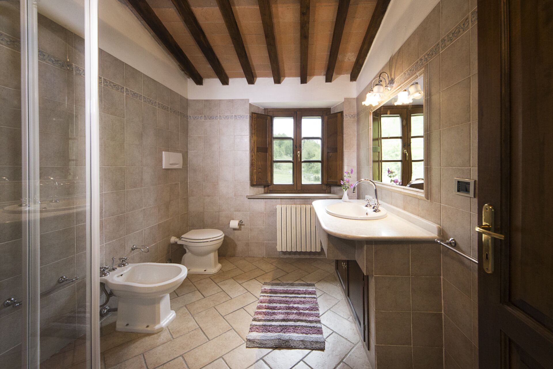A bathroom with a bidet, toilet, and sink, wooden beams on the ceiling, and a window with a view of greenery.