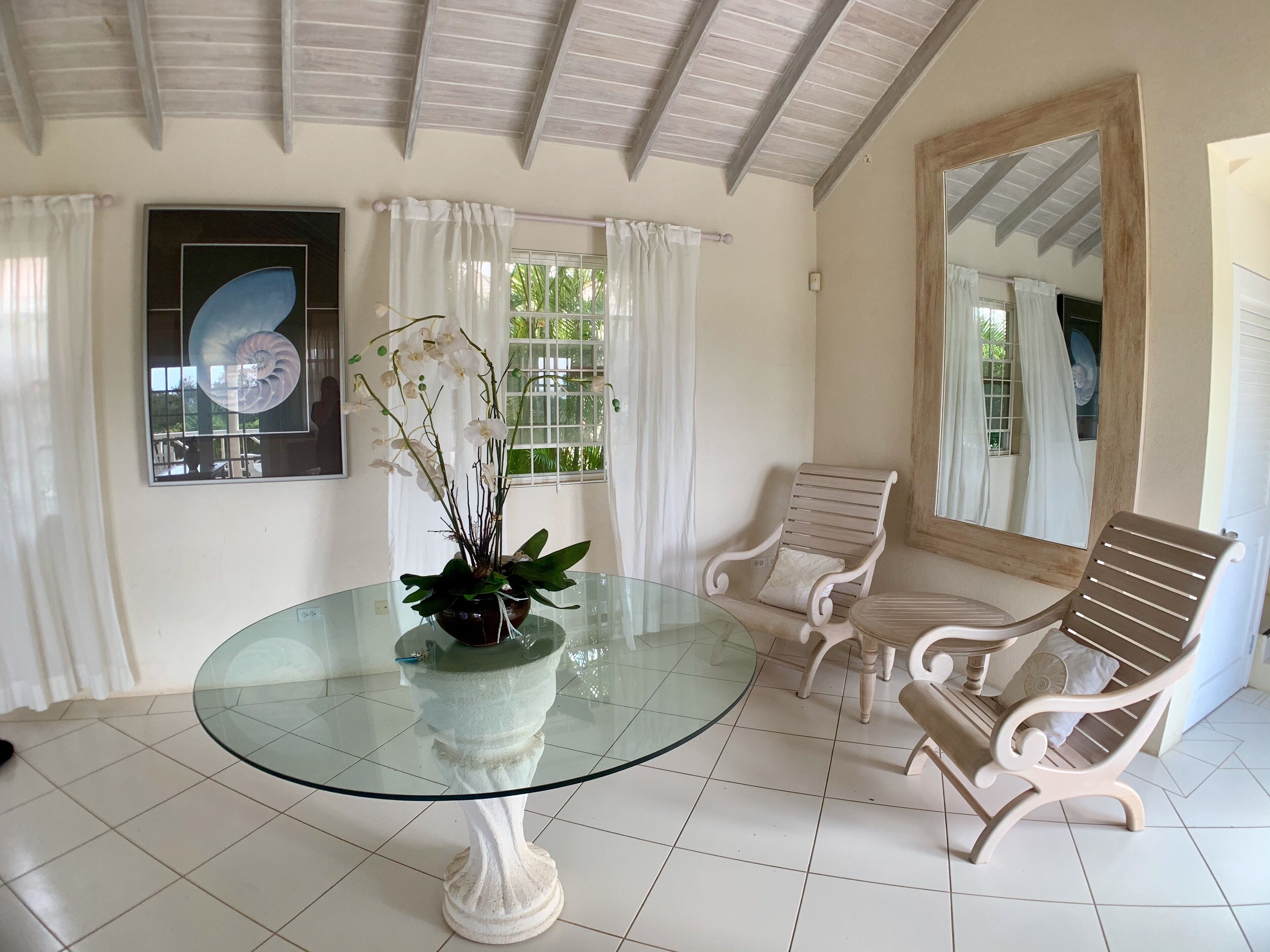 Un coin repas avec une table en verre, des chaises blanches, une plante en pot et un grand miroir.