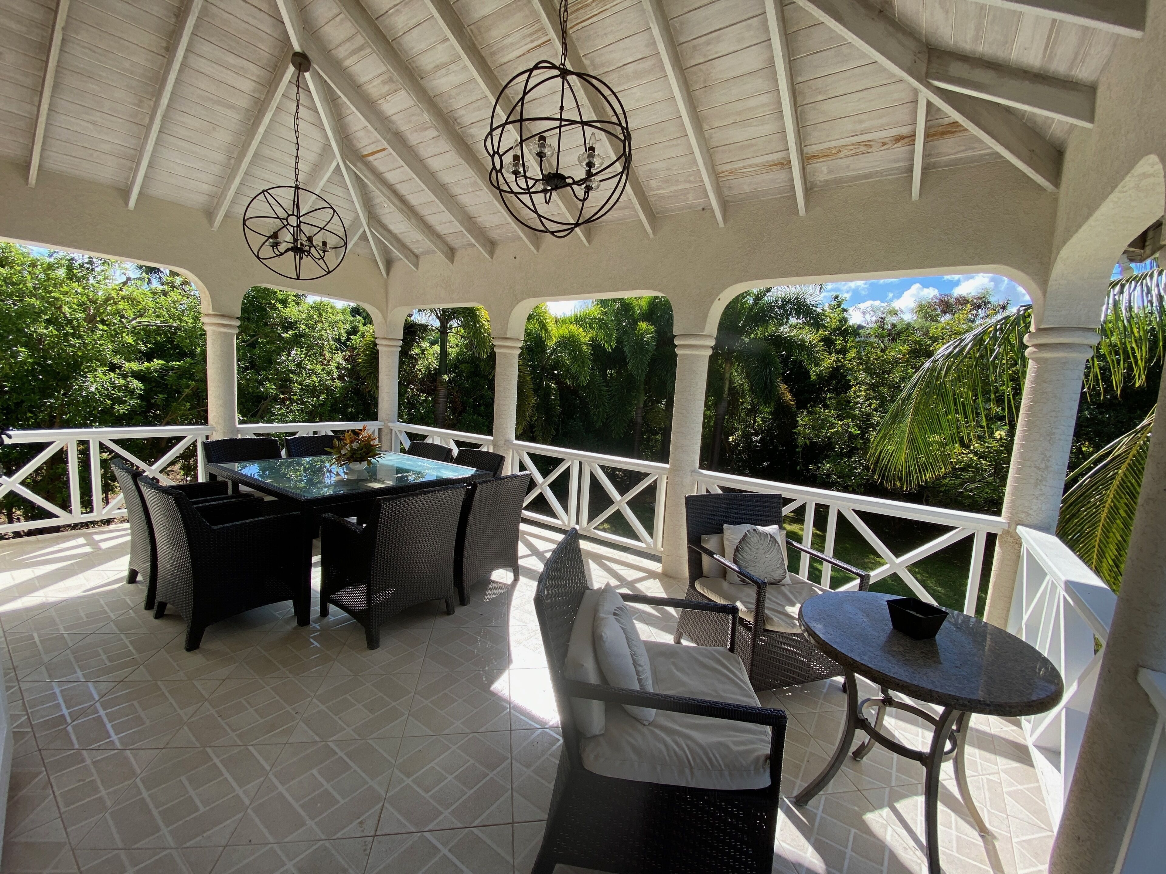 Une terrasse couverte avec une table et des chaises à plateau en verre, entourée d’une balustrade blanche.