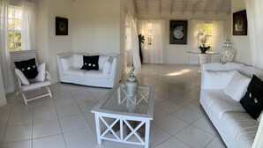 Un salon spacieux, meublé de blanc, avec une table basse en verre et un plafond haut aux poutres apparentes.