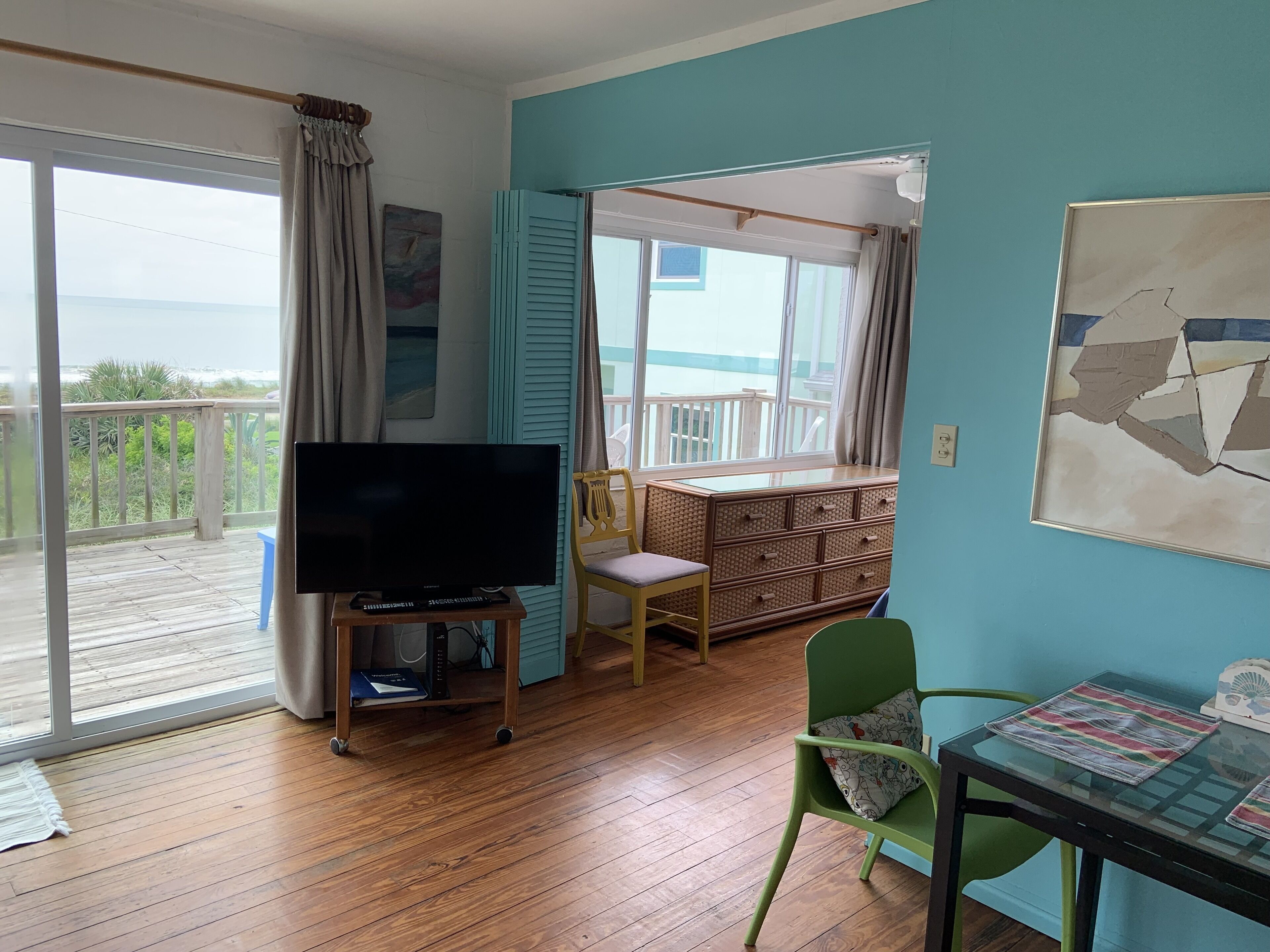 Una habitación con suelo de madera, un televisor sobre un mueble, una silla verde, una mesa con superficie de vidrio y vistas al mar a través de una puerta corrediza.