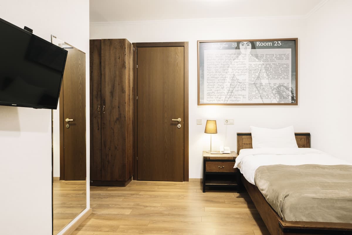 A hotel room with a wooden wardrobe, a bed with a beige blanket, a nightstand with a lamp, and a framed text on the wall.