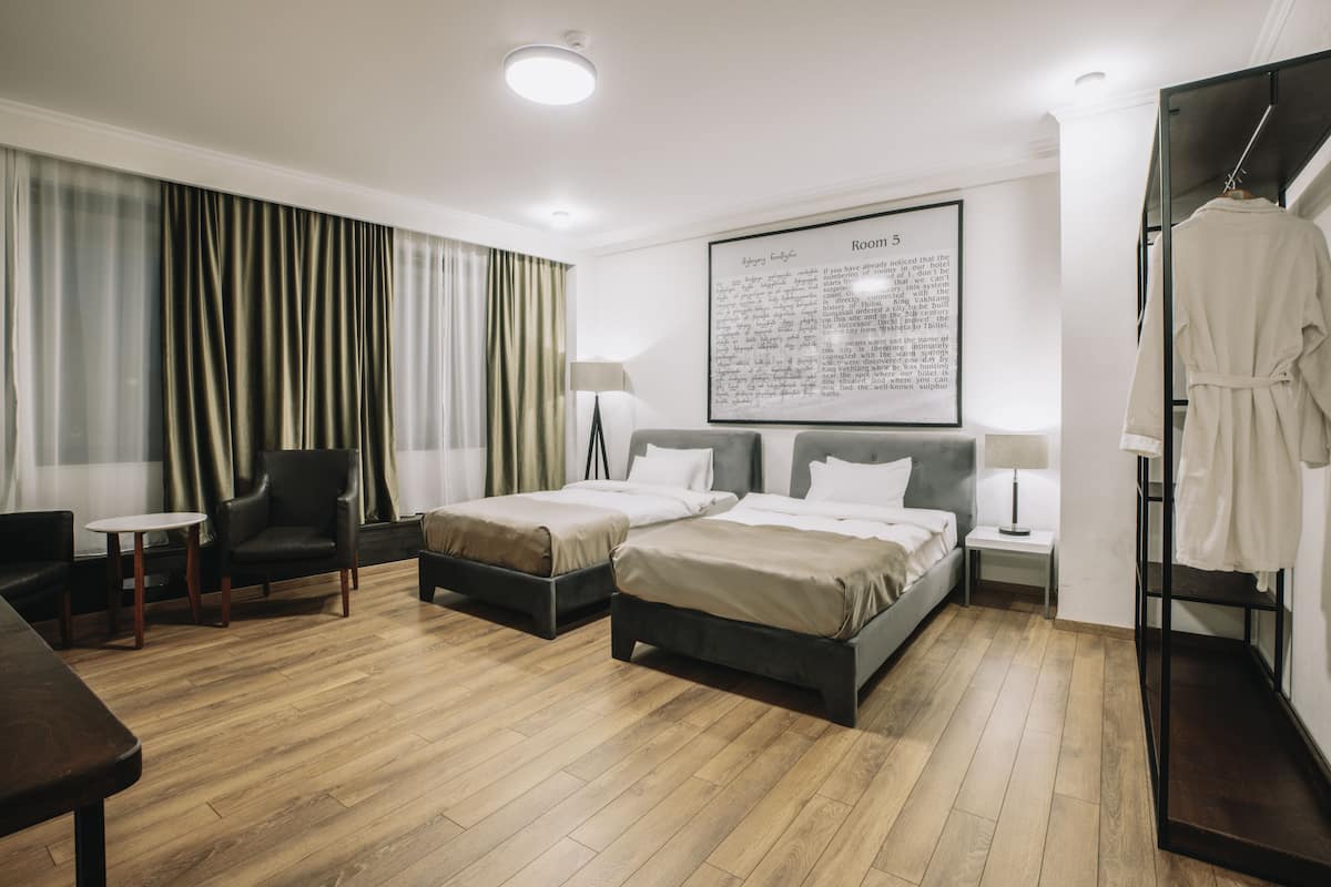 A hotel room with two beds, a chair, a table, a robe rack, and a framed text on the wall.