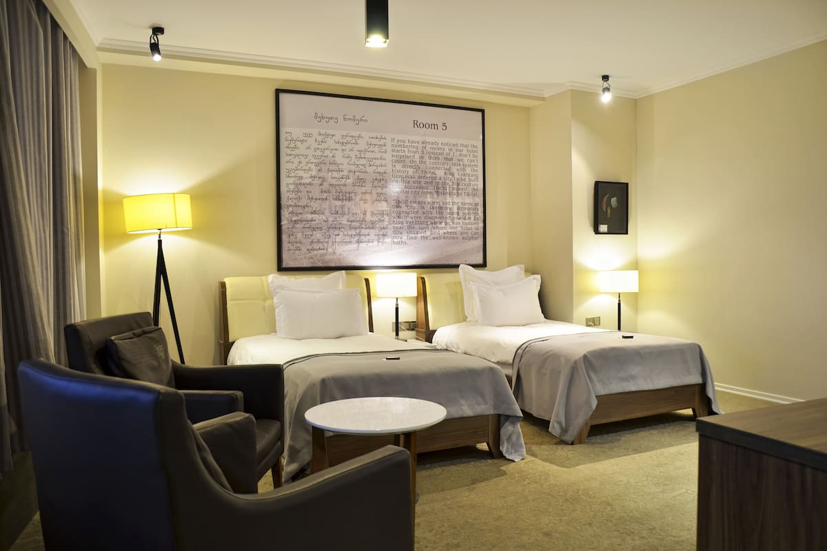 A hotel room with two beds, a desk, a chair, a lamp, and a large framed text on the wall.