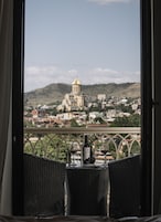 Un balcón con mobiliario de mimbre, una botella de vino y dos copas. Al fondo, se ve una vista de la ciudad con un edificio destacado con cúpula dorada.