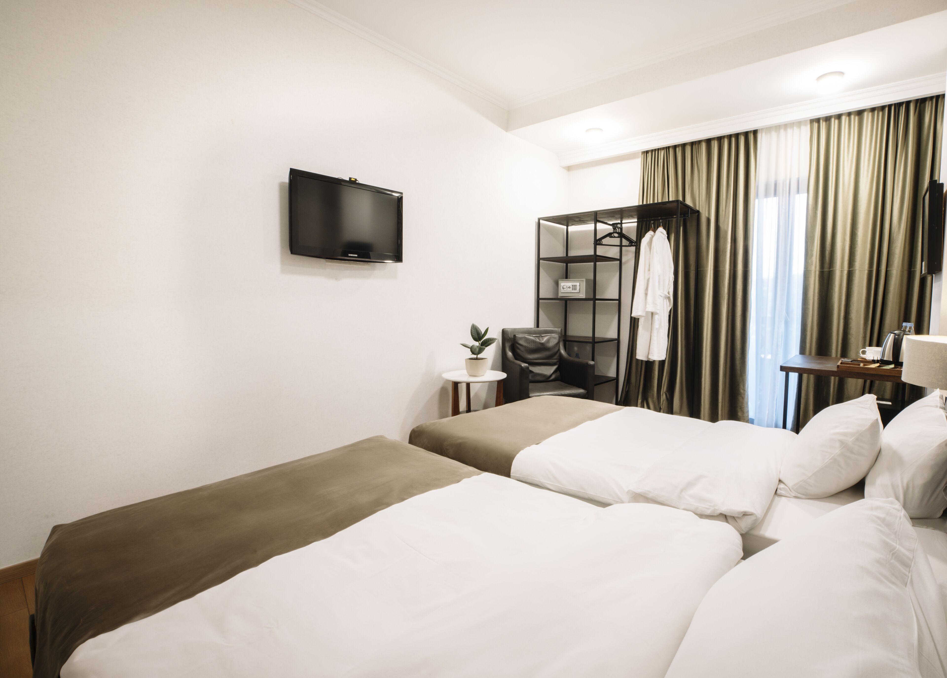 A hotel room with two beds, a TV, a desk, a chair, a shelf with clothes, and a window with curtains.
