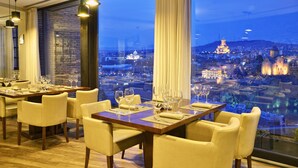 A dining area with tables set for six, large windows with curtains, and a view of a cityscape at dusk. - Citadel Narikala Hotel (Tbilisi)