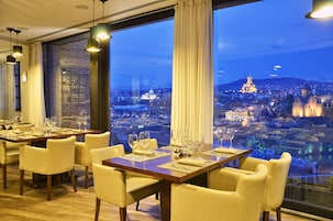 Comedor con mesas dispuestas para seis, ventanas amplias con cortinas y vistas a un paisaje urbano al atardecer.