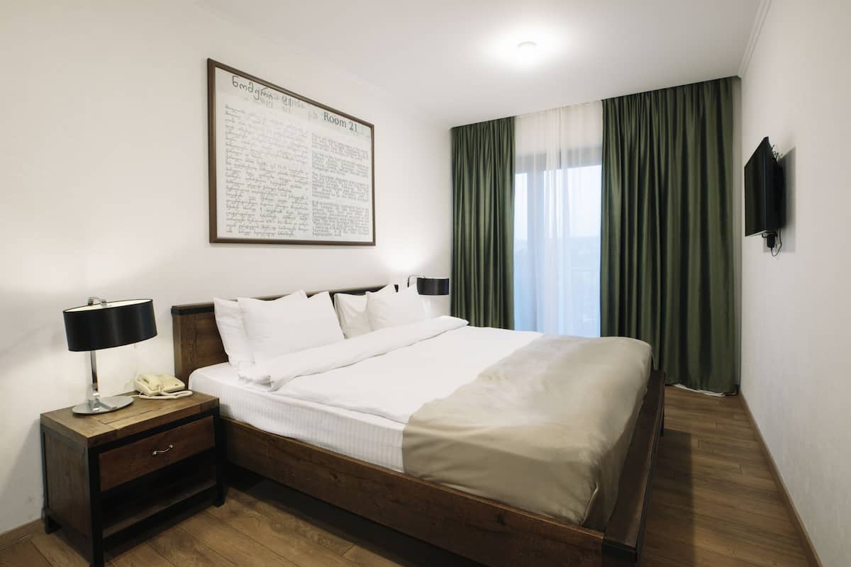 A neatly arranged hotel room with a large bed, a bedside table with a lamp, a mounted TV, and a framed document on the wall.