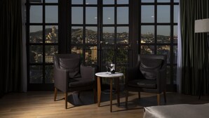 A hotel room with a large window offering a city view, two leather chairs, a small round table with a wine glass, and a lamp on the right. - Citadel Narikala Hotel (Tbilisi)
