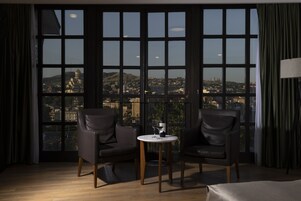 Habitación de hotel con un ventanal grande que ofrece vistas a la ciudad, dos sillones de cuero, una mesita redonda con una copa de vino y una lámpara a la derecha.