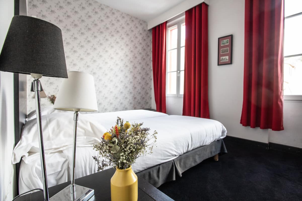 Een hotelkamer met een bed, een bureau met een lamp, een vaas met bloemen en rode gordijnen.