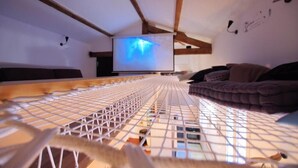 A room with a wooden ceiling, a projector screen displaying a blue image, a bed with a white net, and a dark-colored sofa. - Hostel les Cordées Liées (Albi)
