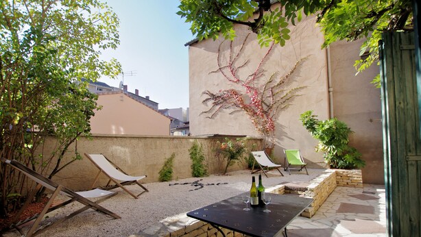 A patio area with two lounge chairs, a table with two wine glasses and bottles, and a decorative vine on the wall. - Hostel les Cordées Liées (Albi)