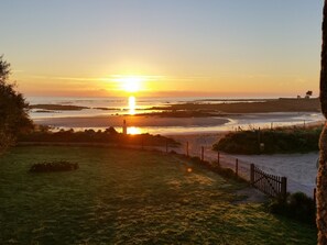 A sunset view from a grassy area with a fence and a path leading to the beach.