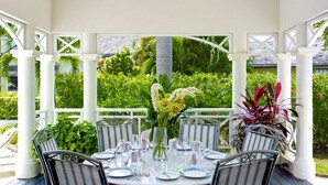 Un espace extérieur abrité, aménagé pour les repas, avec une table ronde pour quatre personnes, entourée de colonnes et de chaises blanches.
