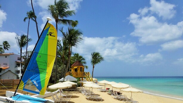Une plage avec des chaises longues et des parasols, un voilier et une petite construction en bois.