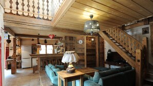Interior rústico com vigas de madeira, escada, cozinha e área de estar com mesa e luminária.