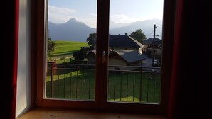 A room with a large window overlooking a green landscape, a mountain in the distance, and a view of a street with buildings.