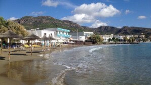 A beach with umbrellas, a building with a blue facade, and a mountain in the background. - Rejuvenate family & friends at Stunning Villa Estia - close to beach & tavernas. (Makry Gialos)