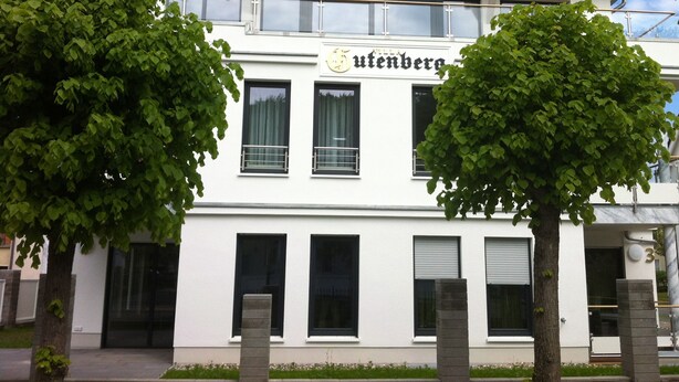 Un edifici de color blanc amb el nom "Mittelguttenberg" a la façana, amb un disseny simètric, grans finestres i un balcó.