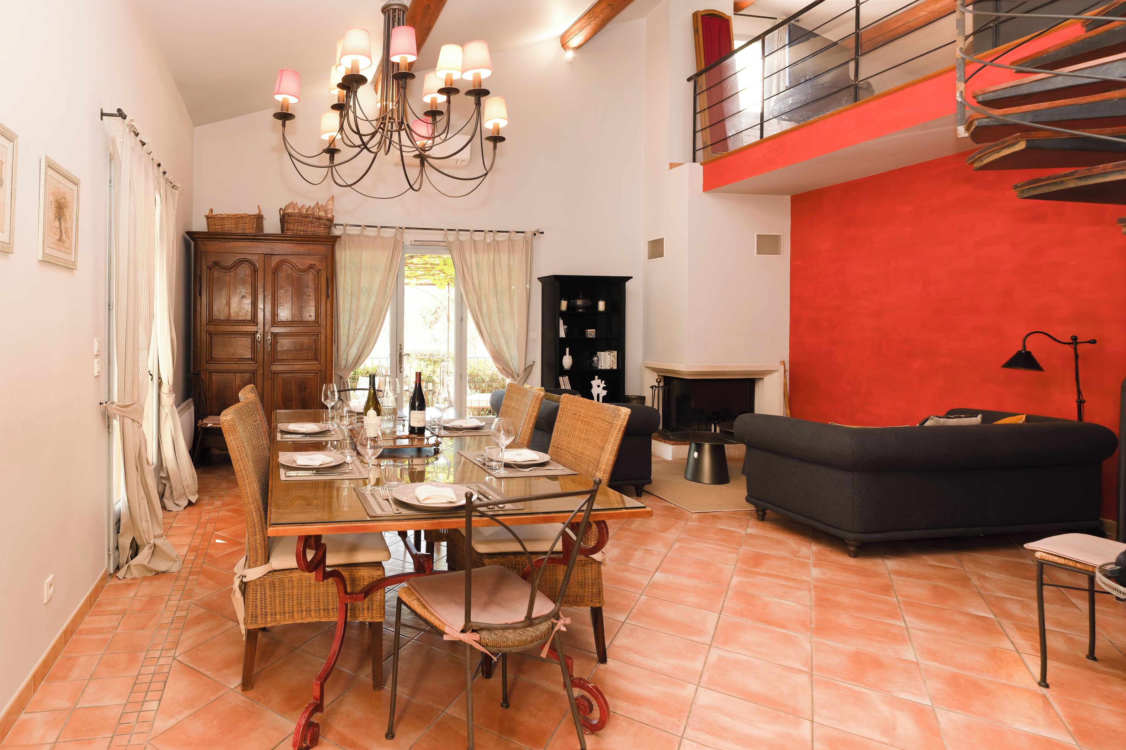 A dining area with a wooden table set for four, surrounded by wicker chairs. A chandelier hangs above, and a black sofa is visible in the background.