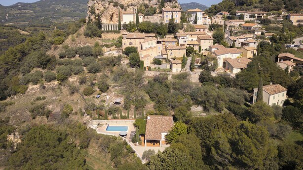 Un village perché sur une colline, avec des bâtiments traditionnels en pierre, une formation rocheuse remarquable et une piscine.
