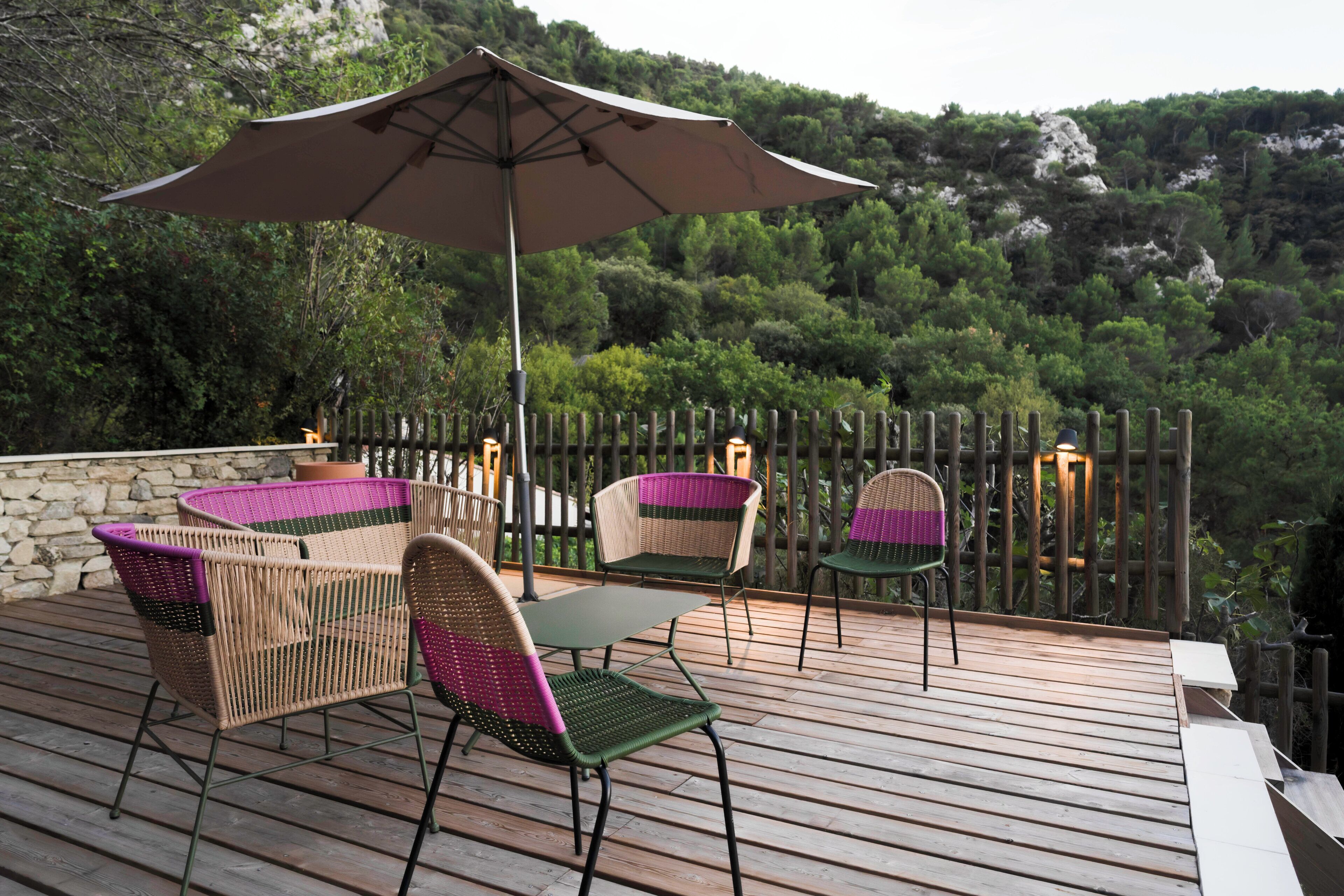 A wooden deck with wicker chairs and a table, surrounded by a stone wall and a wooden fence, overlooking a forested hillside.